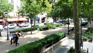 Rua com duas calçadas, sendo uma elas bem ampla, com bancos, alguns transeuntes, jardins e diversas árvores