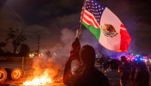 TOPSHOT – Uma manifestante agita uma bandeira combinando os símbolos dos Estados Unidos e do México enquanto forças de segurança entram em confronto com manifestantes durante um protesto após operações federais de imigração, no bairro de Compton, em Los Angeles, Califórnia, em 7 de junho de 2025. O presidente dos EUA, Donald Trump, mobilizou 2.000 soldados neste dia para conter os protestos crescentes contra as ações de repressão migratória na região de Los Angeles — medida que o governador do estado classificou como "deliberadamente provocativa". Agentes federais entraram em confronto com multidões enfurecidas em um subúrbio de Los Angeles, enquanto os protestos chegavam à segunda noite no sábado, com uso de granadas de efeito moral e o bloqueio de parte de uma rodovia durante operações contra imigrantes indocumentados, segundo relatos. (Foto: Ringo Chiu / AFP)
